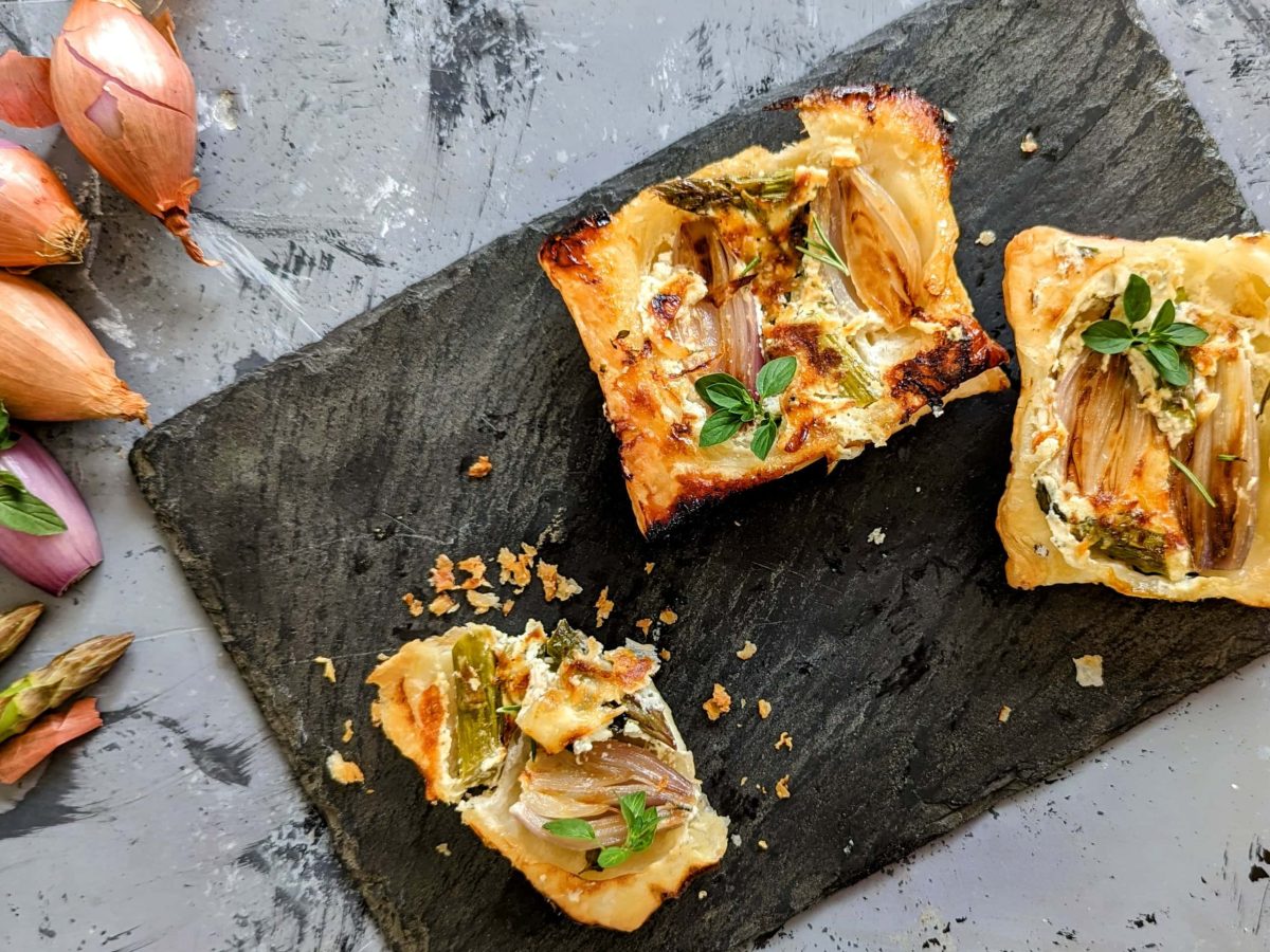 Eine Schieferplatte mit ein paar Stücken der Upside-Down-Tarte direkt von oben fotografiert. Alles auf einem Steintisch. Daneben noch grüner Spargel und Schalotten.