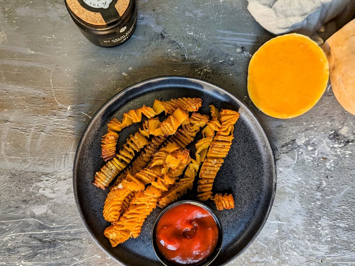 Ein schwarzer Teller mit den Kürbis-Akkordeon-Pommes und Ketchup auf einem grauen Steintisch. Alles direkt von oben fotografiert.