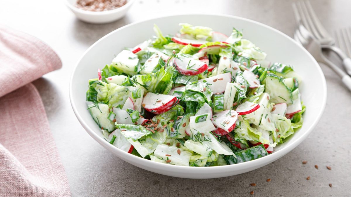 Draufsicht schräg: Eine weiße Schüssel Radieschen-Gurken-Salat auf hellem Untergrund. Daneben liegt ein Besteck.