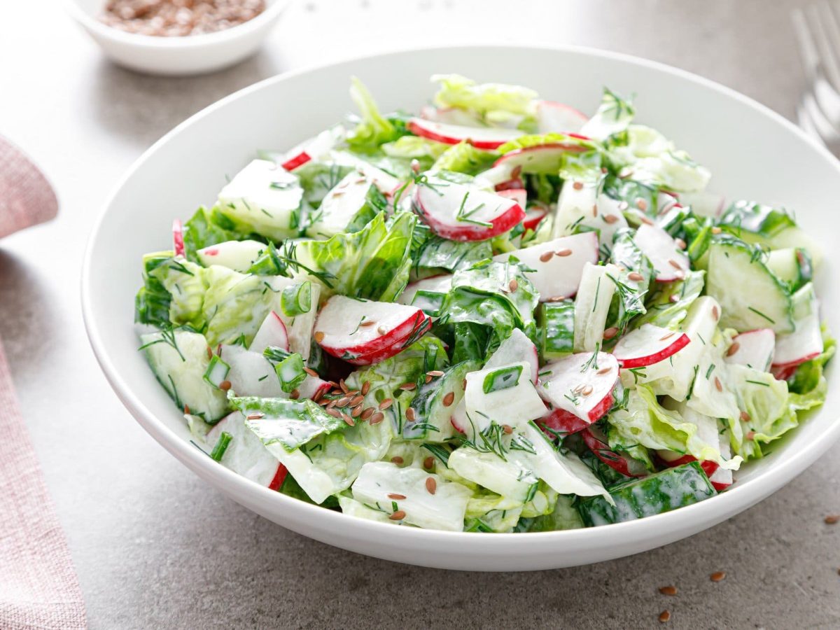 Draufsicht schräg: Eine weiße Schüssel Radieschen-Gurken-Salat auf hellem Untergrund. Daneben liegt ein Besteck.