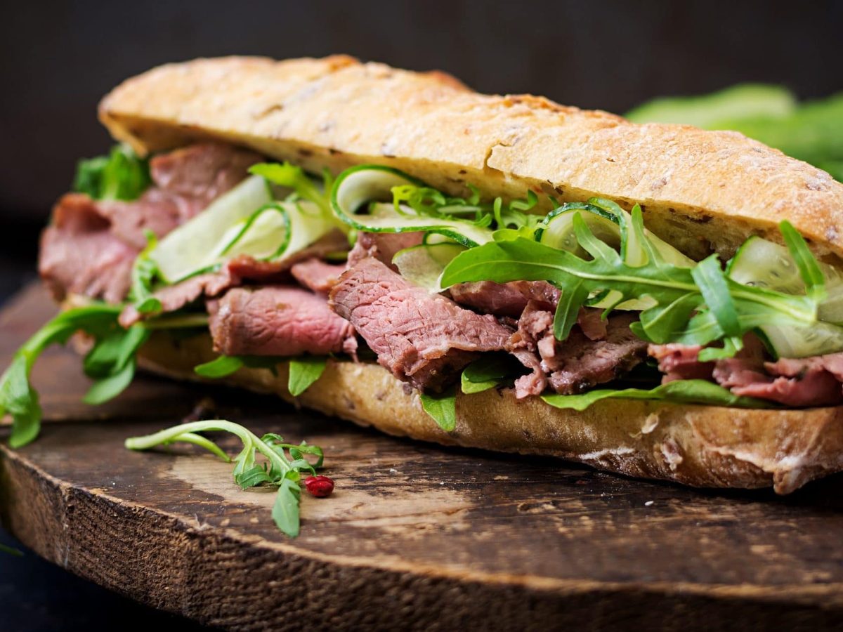 Frontal: ein Roastbeef-Gurken-Sandwich vor dunklem Hintergrund auf einem runden Holztisch.