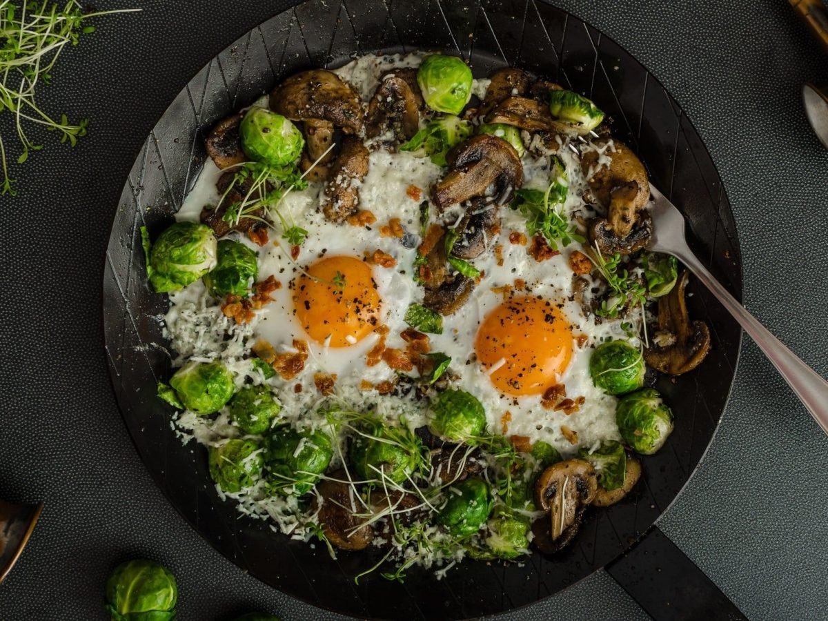 Draufsicht: eine Rosenkohl-Champignon-Pfanne mit zwei Spiegeleiern auf einem dunklen Untergrund. Daneben liegen Sprossen und ein Besteck.