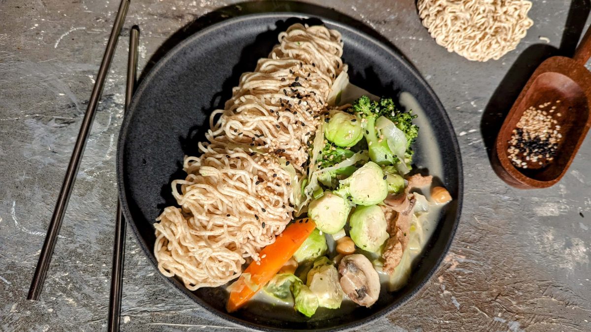 Ein schwarzer Teller mit dem Rosenkohl-Curry sowie Mie-Nudeln auf einem Steintisch von oben fotografiert. Daneben Sesam, ungekochte Mie-Nudeln und zwei Essstäbchen.