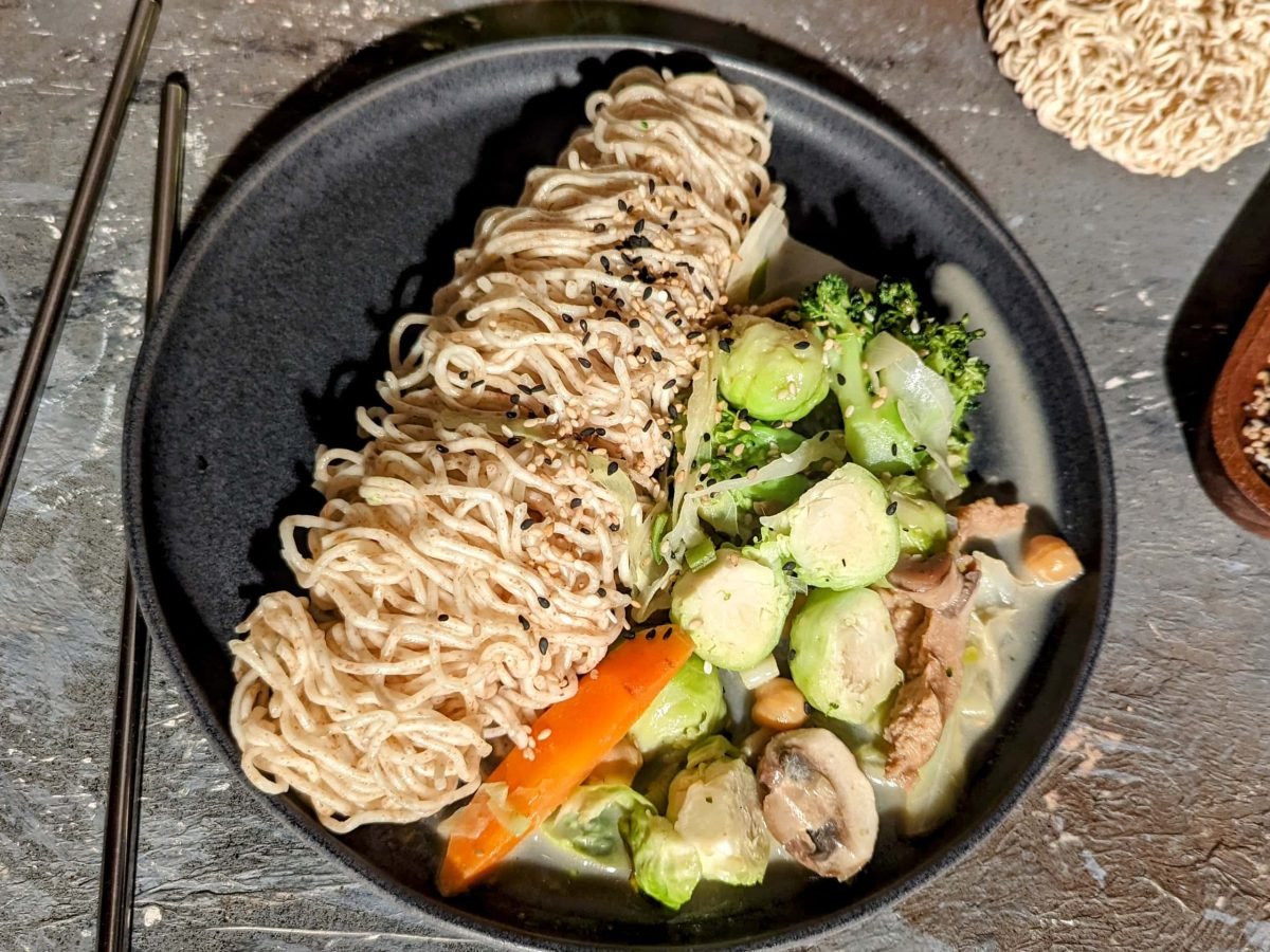 Ein schwarzer Teller mit dem Rosenkohl-Curry sowie Mie-Nudeln auf einem Steintisch von oben fotografiert. Daneben Sesam, ungekochte Mie-Nudeln und zwei Essstäbchen.