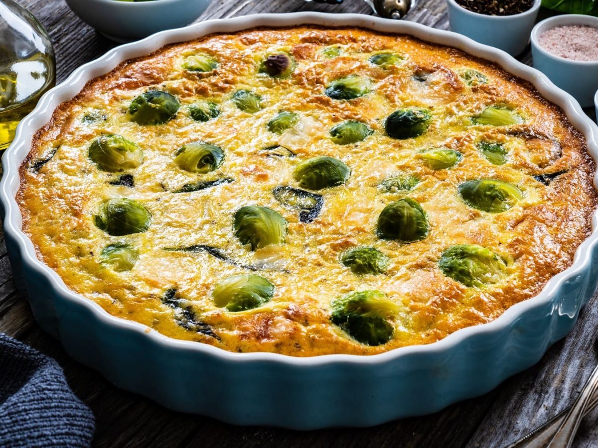 Eine blaue Auflaufform mit der Rosenkohl-Quiche auf einem dunklen Holztisch von schräg oben fotografiert.