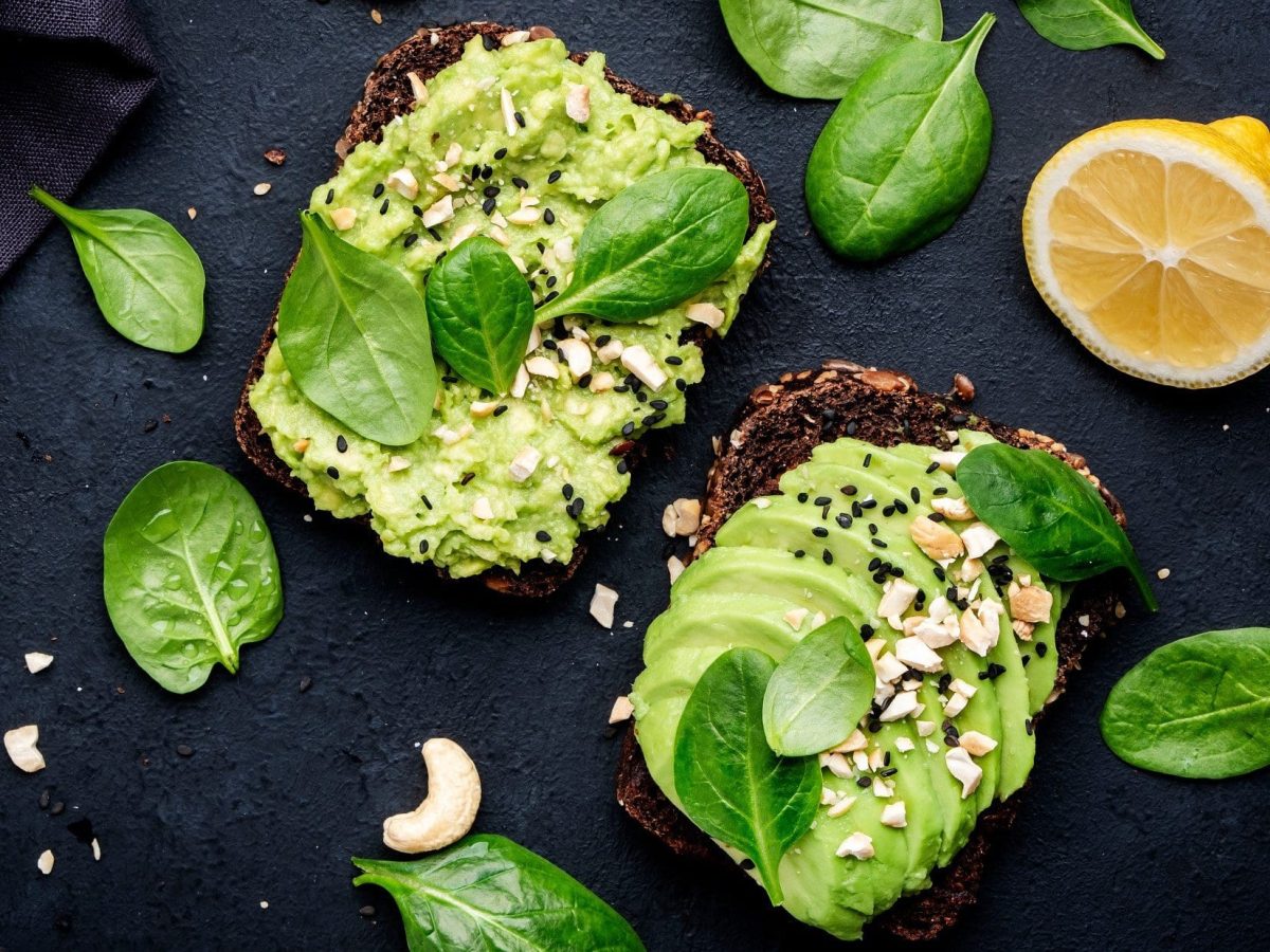 Draufsicht: Schwarzer Untergrund, zwei Sandwich mit Avocado-Cashew-Creme, Spinatblätter, halbierte Zitrone.