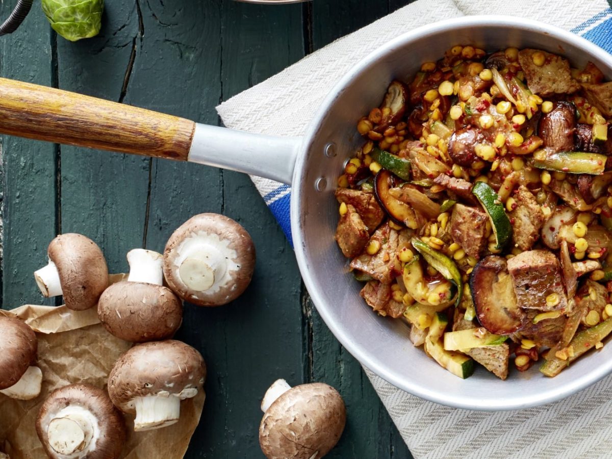 Eine Schnitzel-Gemüse-Pfanne mit Linsen auf einem Geschirrtuch auf einem dunkelblauen Hintergrund. Links unten Champignons und eine Flasche Öl.