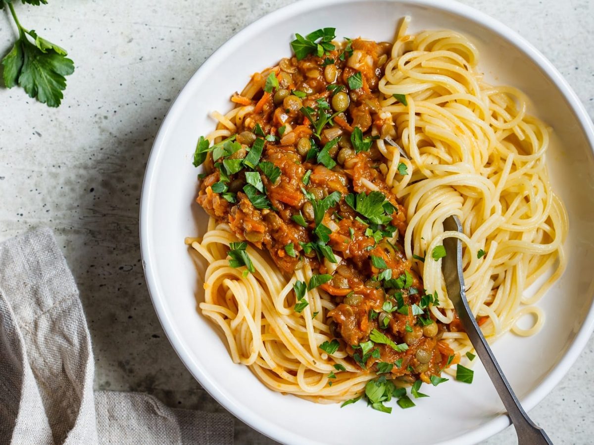 Ein weißer Teller mit Spaghetti und Linsen-Bolognese auf einem grauen Steintisch direkt von Oben fotografiert. Links vom Bild außerdem, Petersilie und ein graues Tuch. Rechts im Bild eine silberne Gabel, die in den Teller ragt.