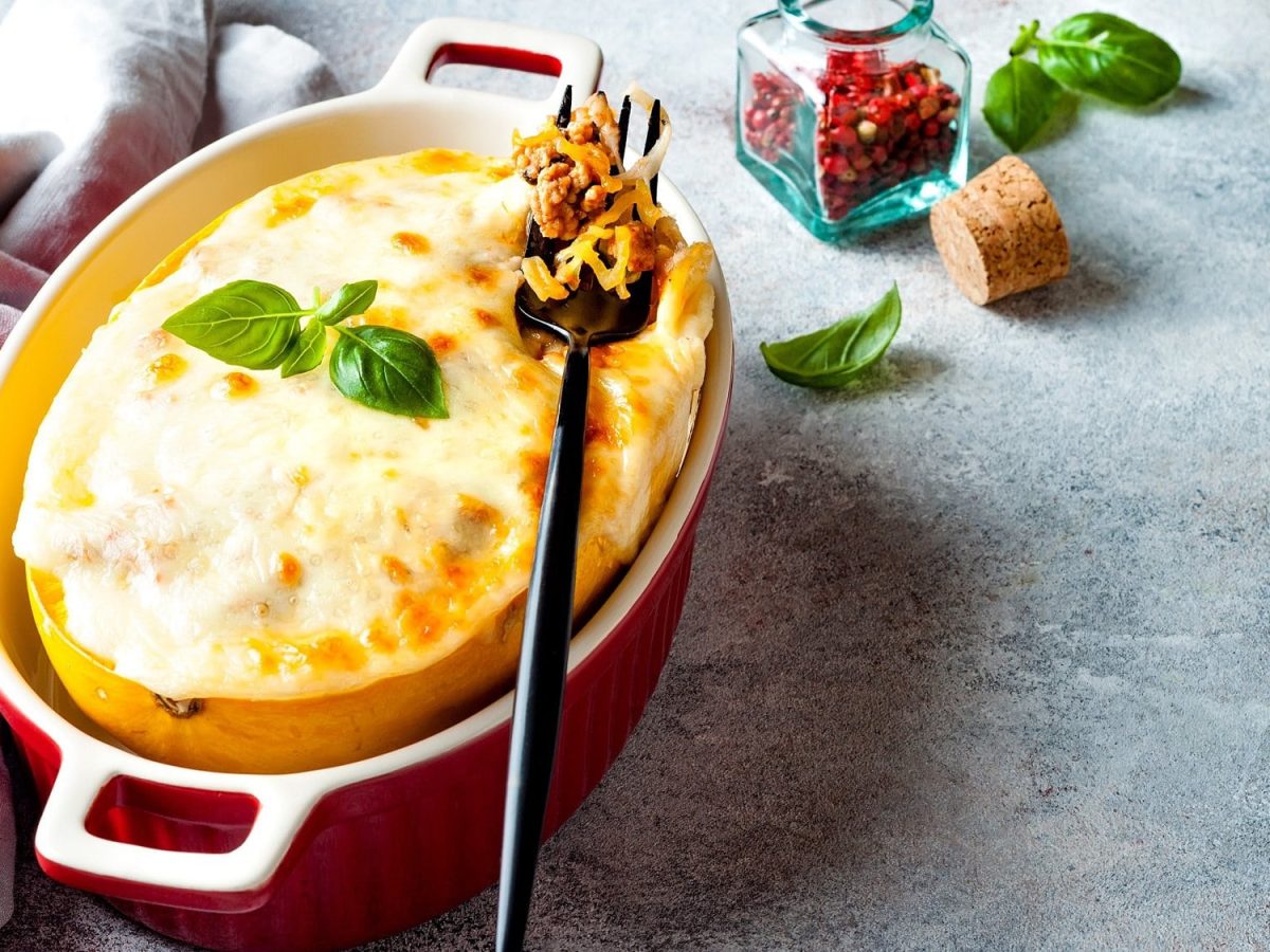 Ein halber Spaghettikürbis Elsässer Art in einer roten Auflaufform, mit Käse überbacken und Basilikum garniert, darauf eine Gabel, drumherum etwas frischer Basilikum und ein Glas mit rotem Pfeffer.