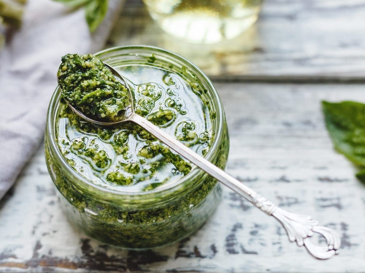 In einem Glas ist eine Spinat-Pesto. Auf dem Glas liegt ein Löffel. Neben dem Glas liegt ein Blass Spinat.