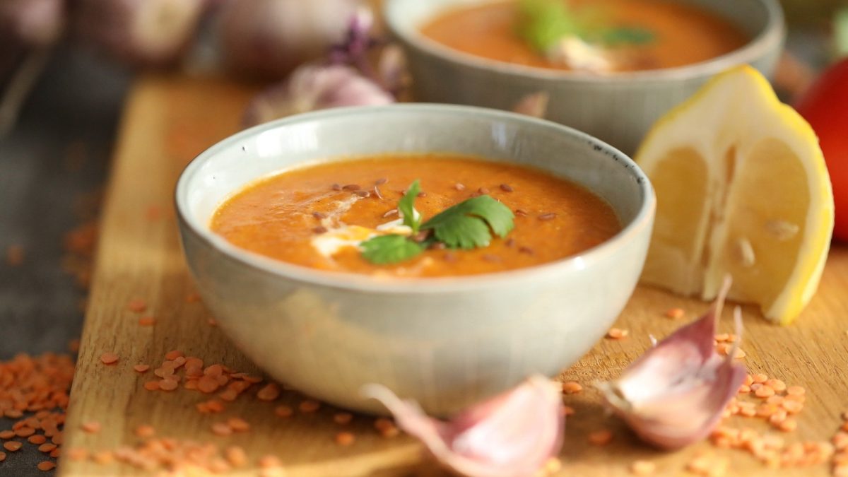 Zwei Schüsseln Süßkartoffelsuppe mit Linsen hintereinander auf einem Brett, daneben rohe Linsen, Knoblauchzehen und eine geviertelte Zitrone.