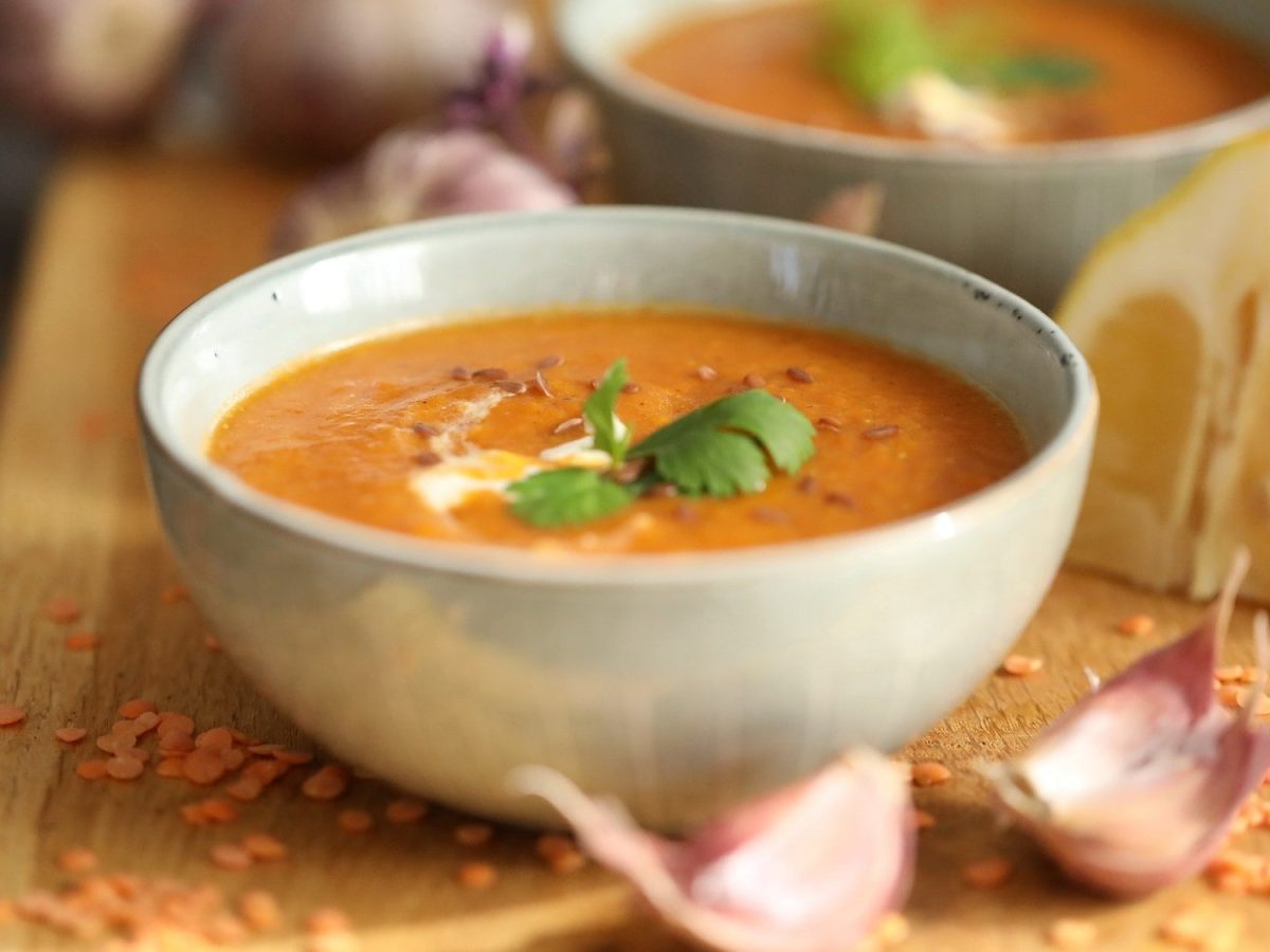 Zwei Schüsseln Süßkartoffelsuppe mit Linsen hintereinander auf einem Brett, daneben rohe Linsen, Knoblauchzehen und eine geviertelte Zitrone.