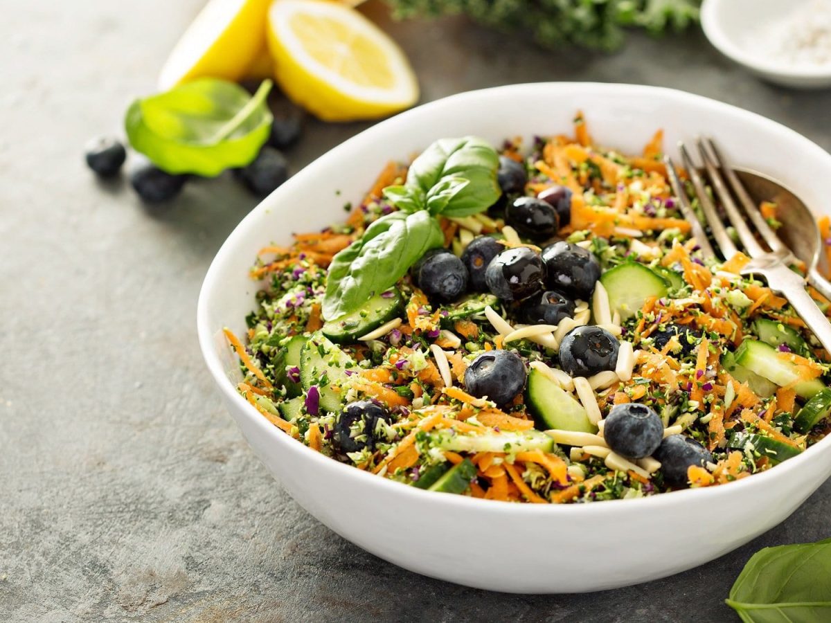 Eine Schüssel Superfood-Salat mit geraspelter Möhre, zerkleinertem Brokkoli, Heidelbeeren, Gurke, darin Besteck, daneben eine aufgeschnittene Zitrone.