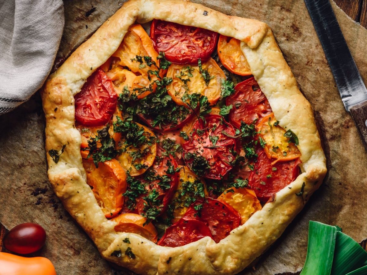 Eine Vollkorn-Tomaten-Galette auf Backpapier auf hölzernem Hintergrund. Rechts ein Messer, rechts oben eine kleine Schüssel mit Kräutern. Links ein graues Geschirrtuch und Tomaten.