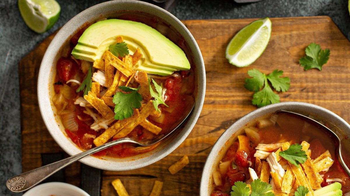 Zwei Schüsseln Tortilla-Suppe mit Koriander, Avocadoscheiben und Tortillastreifen garniert, darin ein Löffel, drumherum Limettenspalten. Draufsicht.