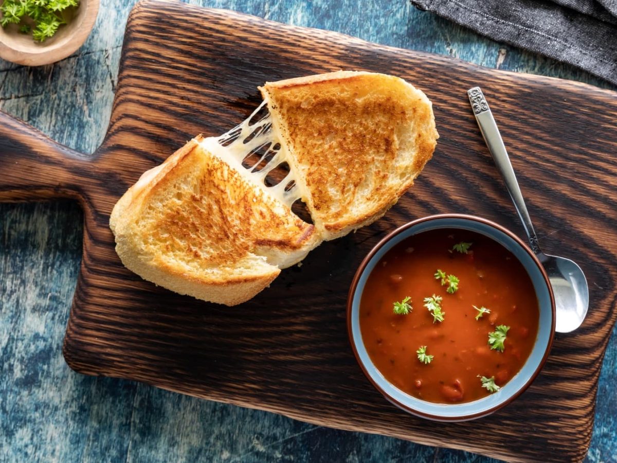 Ein Toast mit veganem Mozzarella gefüllt auf einem hölzernen Schneidebrett auf einem dunkelgrauen Hintergrund. Auch noch auf dem Brett eine kleine Tomatensuppe und ein Löffel. Links oben ein Schüsselchen mit Petersilie.