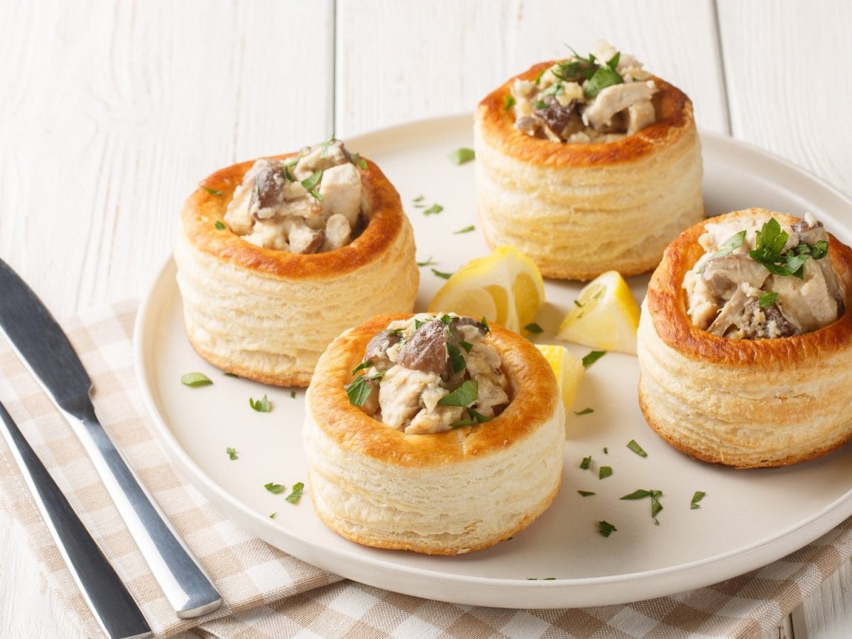 4 Vol-au-vent mit Pilzen auf einem weißen Teller, dazwischen Zitronenspalten, daneben Messer und Gabel sowie eine Serviette.