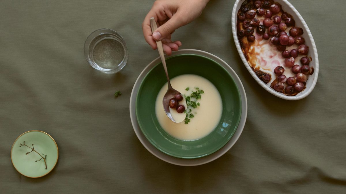 Eine Schüssel Wintersuppe, eine Hand legt mit einem Esslöffel darauf karamellisierte Trauben ab. Daneben eine Auflaufform mit den Trauben. Draufsicht.