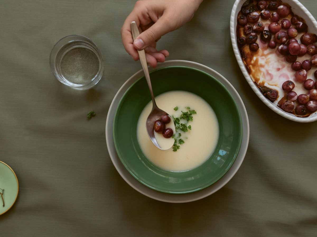 Eine Schüssel Wintersuppe, eine Hand legt mit einem Esslöffel darauf karamellisierte Trauben ab. Daneben eine Auflaufform mit den Trauben. Draufsicht.