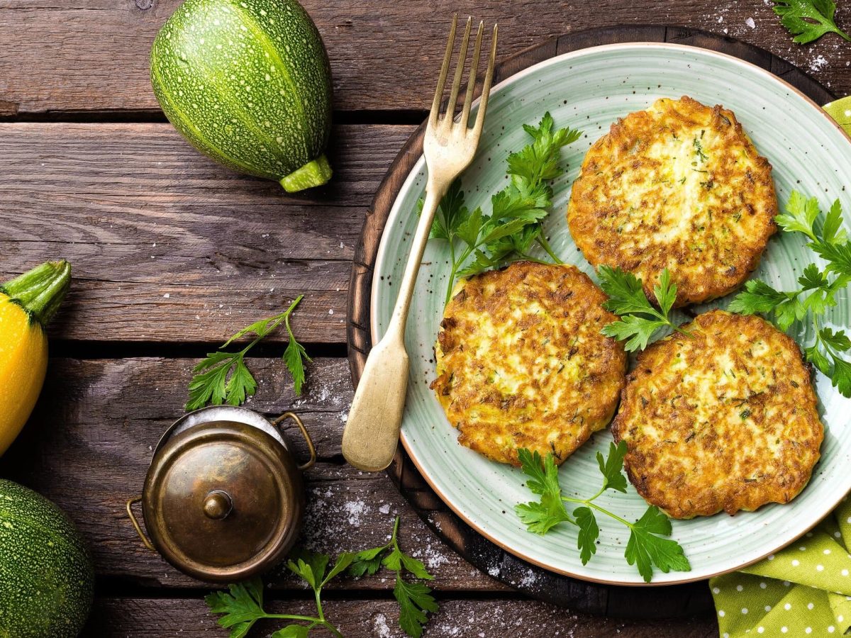 Draufsicht: Auf einem Holztisch steht ein Teller mit Zucchini-Puffer mit Hüttenkäse. Drum herum liegen Zucchini und ein gelbes Tuch.