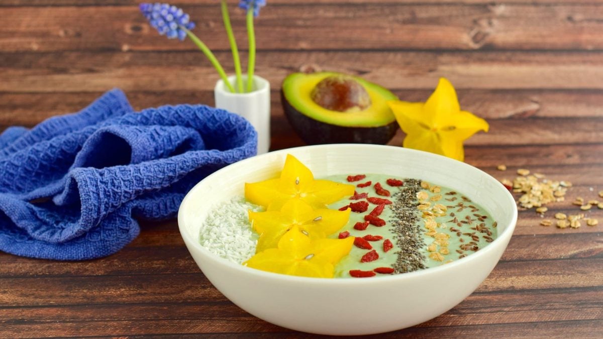 Draufsicht, schräg: eine Avocado-Bowl mit Goji-Beeren auf Holzboden. Drum herum steht eine Vase mit blauen Blumen. Daneben liegen ein blaues Tuch, eine halbe Avocado und eine Sternfrucht.