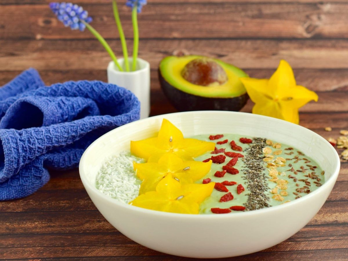 Draufsicht, schräg: eine Avocado-Bowl mit Goji-Beeren auf Holzboden. Drum herum steht eine Vase mit blauen Blumen. Daneben liegen ein blaues Tuch, eine halbe Avocado und eine Sternfrucht.