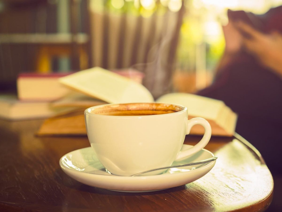 Frontal eine weiße Kaffeetasse mit Untertasse steht in einem Raum auf einem dunklen, runden Holztischchen. Dahinter liegen mehrere Bücher, zum teil aufgeschlagen.