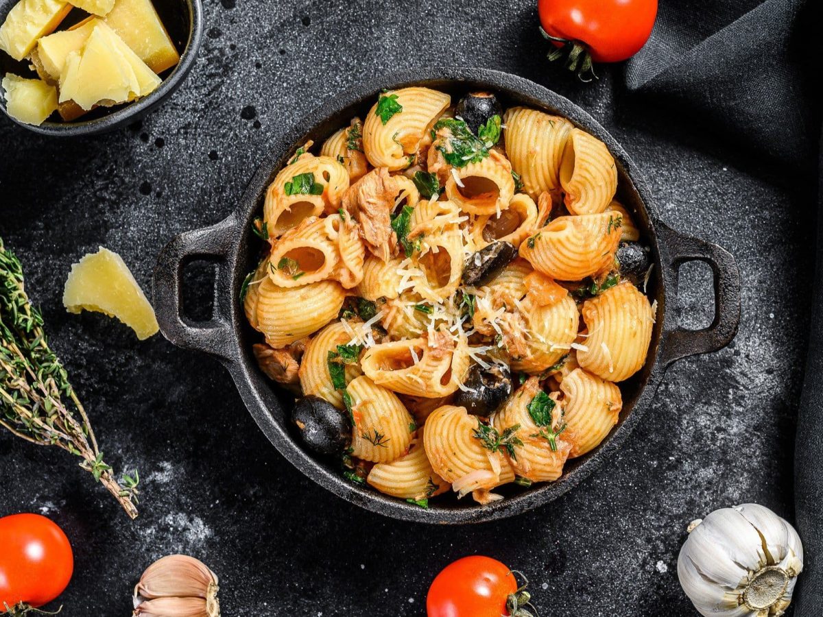 Draufsicht: Eine dunkle Pfanne mit Conchiglie Rigate mit Thunfisch steht auf einem dunklem Untergrund. Drum liegen Tomaten, Knoblauch und Gewürze.