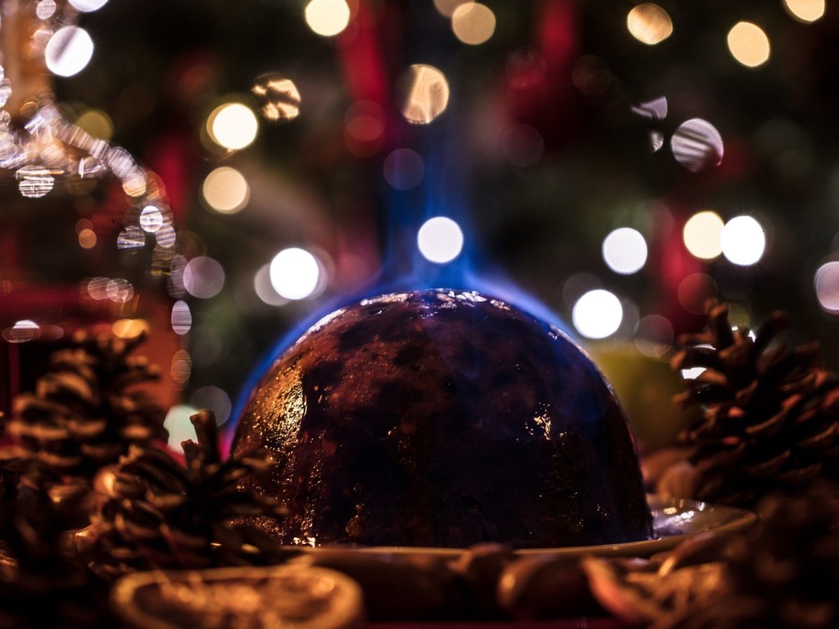 Frontal: Ein britischer Christmas Pudding der flambiert wird. Drum herum ist es weihnachtlich geschmückt.