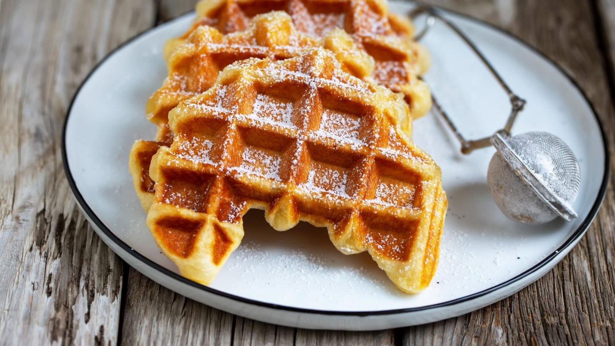 Draufsicht, schräg: Ein weißer Teller mit zwei Croffles und einem Puderzuckersieb auf einem Untergrund aus Holz.