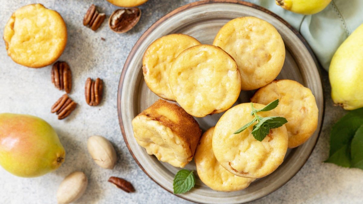 Draufsicht: Mehrere Hüttenkäse-Muffins sowie Nusskerne liegen auf einem Teller und einem hellem Untergrund. Drumherum liegen Birnen.