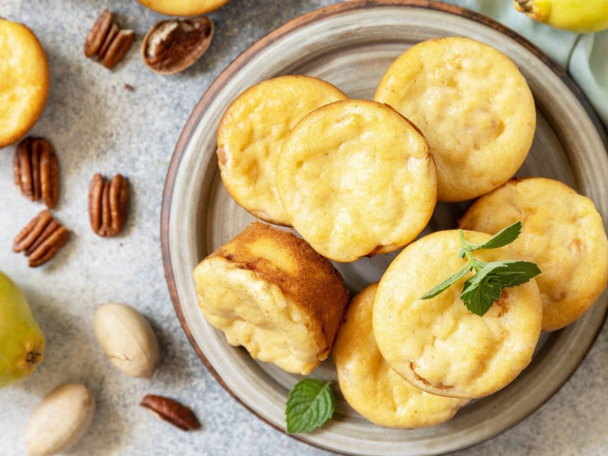 Draufsicht: Mehrere Hüttenkäse-Muffins sowie Nusskerne liegen auf einem Teller und einem hellem Untergrund. Drumherum liegen Birnen.