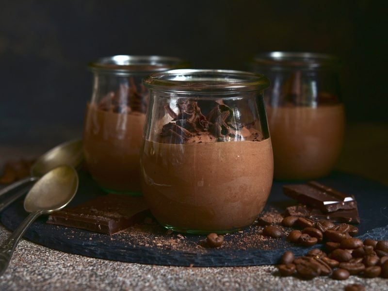 Frontal: Drei Gläser Mousse au Café auf einem Untergrund aus Holz vor einem sehr dunklen Hintergrund.