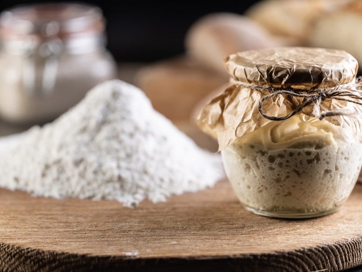 Frontal: Rundes Holzbrett auf dem loses Mehl und ein verschlossenes Glas mit Sauerteig ist. Man macht ihn so haltbar. Im Hintergrund ist ein weiteres Glas.