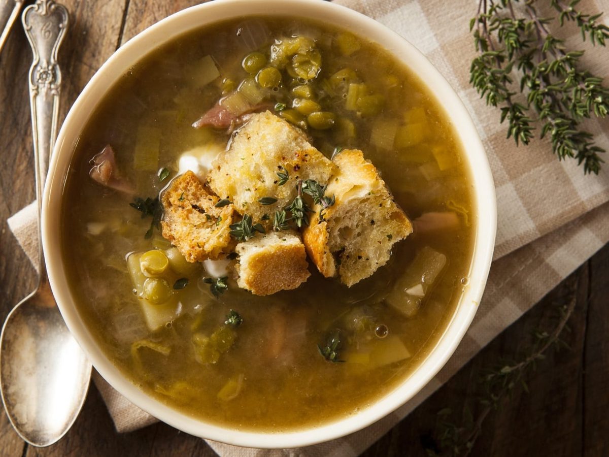 Eine weiße Suppenschale mit der Erbsen-Lauch-Suppe auf einem Holztisch mit frischem Thymian und einer weiß-braun-karierten Tischdecke und zwei Löffeln. Alles von oben fotografiert.