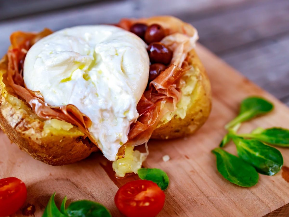 Eine gebackene und halbierte Kartoffel mit Burrata, Schinken und Oliven gefüllt auf einem Holzbrett mit Kirschtomaten und Feldsalat.