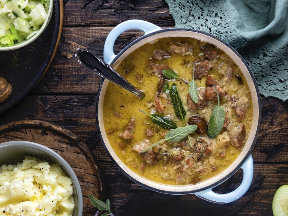 Geschmortes Schweinefleisch mit Äpfeln in einem Schmortopf. Rechts oben ein dunkelgrünes Geschirrtuch, links eine Schüssel mit Kartoffelpüree, links oben eine Schüssel mit Wirsinggemüse. Rechts unten auf dem Bild drei Apfelhälften.