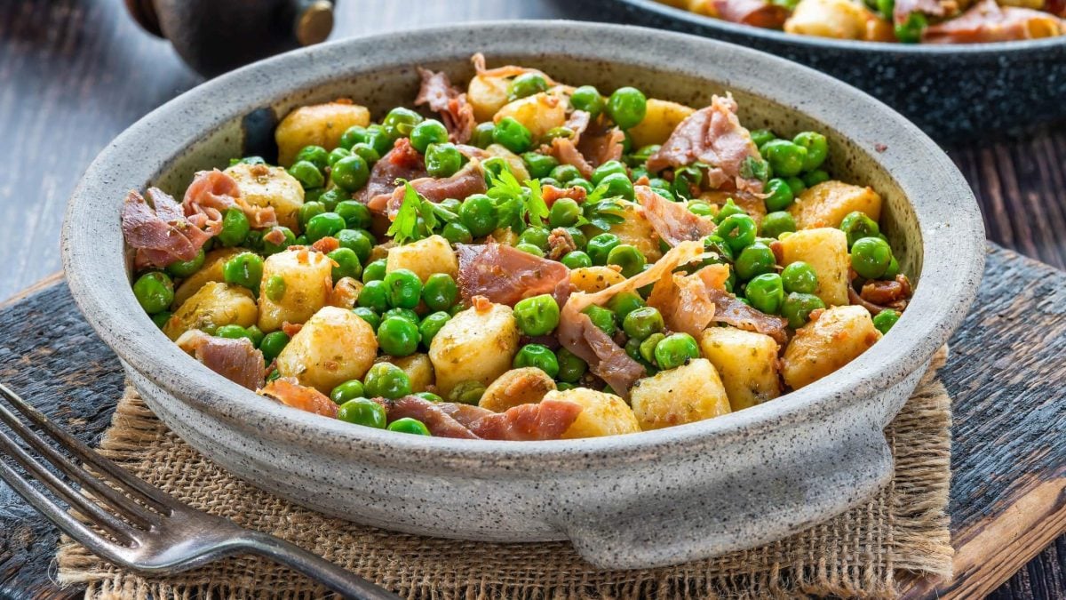 Draufsicht schräg: eine graue Schüssel mit Gnocchi-Pfanne mit Erbsen und Speck steht auf einem groben Leinentuch auf einem Holztisch.