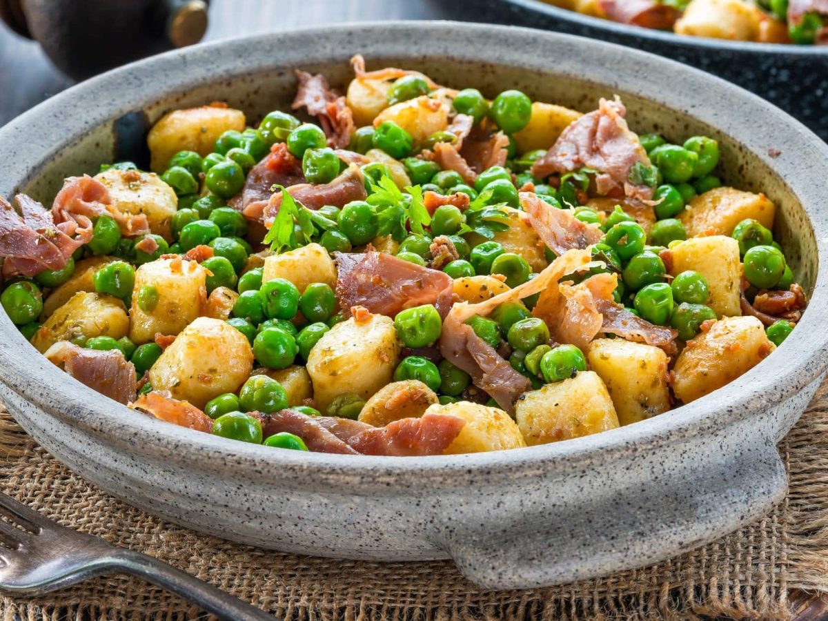 Draufsicht schräg: eine graue Schüssel mit Gnocchi-Pfanne mit Erbsen und Speck steht auf einem groben Leinentuch auf einem Holztisch.