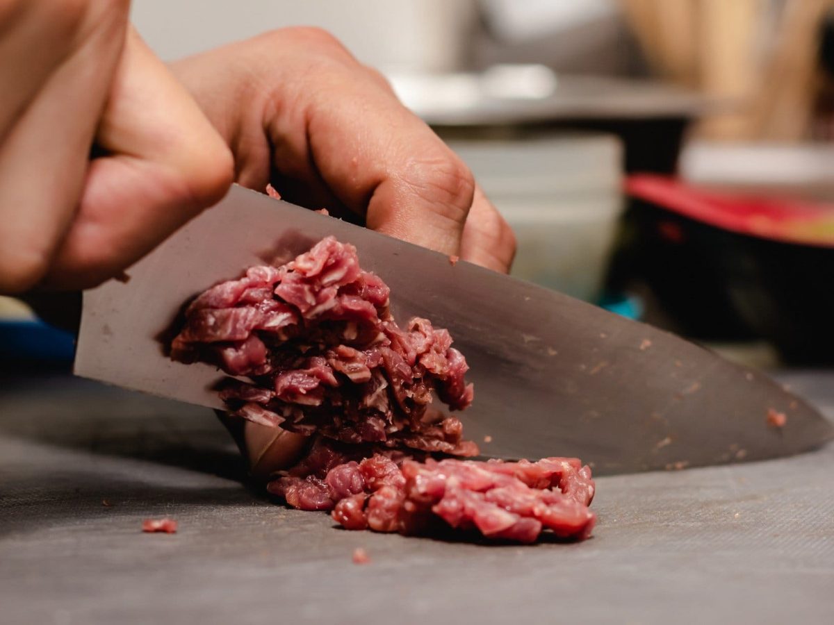 Draufsicht: Ein Mann macht mit einem Küchenmesser Hackfleisch selbst. Man sieht nur die Hand des Mannes.