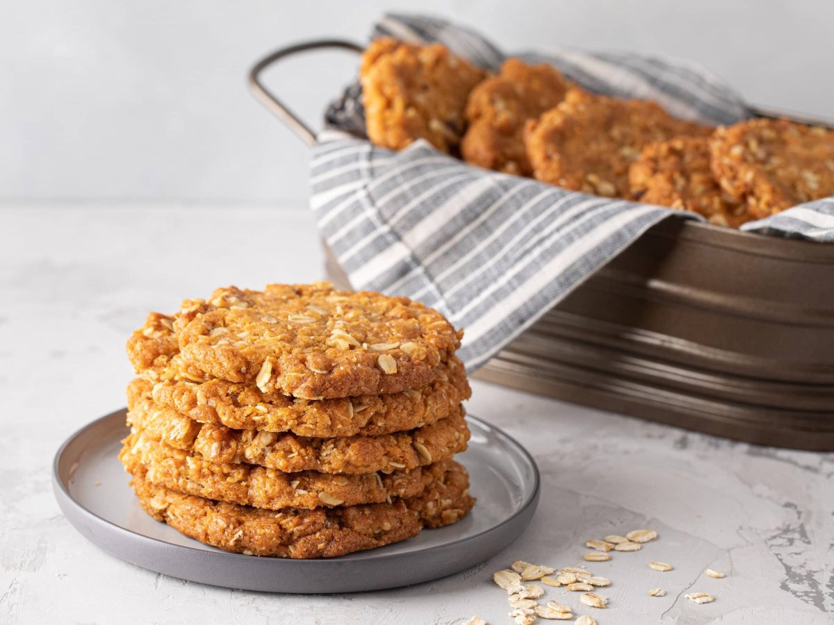 Die Haferflocken-Kokos-Cookies gestapelt auf einem grauen Teller. Im Hintergrund noch mehr Kekse in einer länglichen Backform. Alles von der Seite fotografiert.