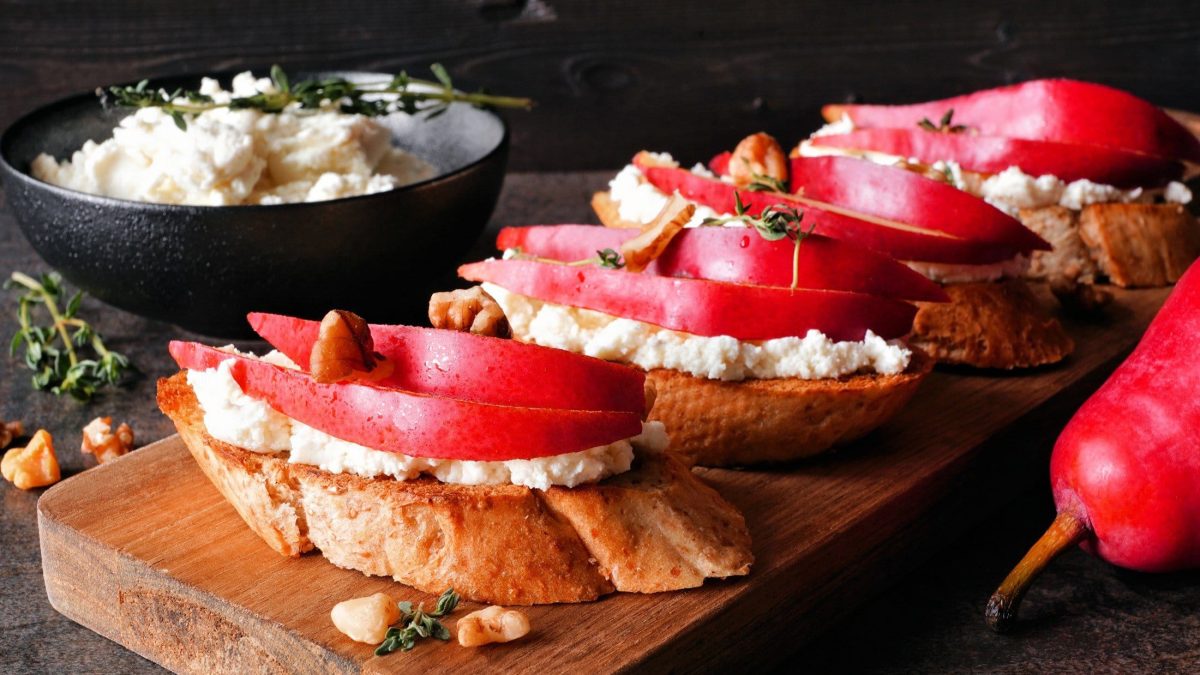 Frontal: Ein Brettchen mit Hüttenkäse-Crostini mit Birne vor einem dunklen Hintergrund.