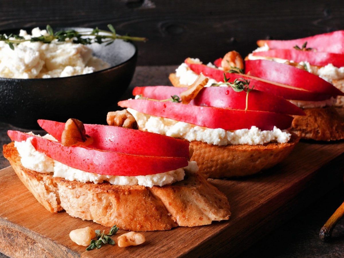 Frontal: Ein Brettchen mit Hüttenkäse-Crostini mit Birne vor einem dunklen Hintergrund.