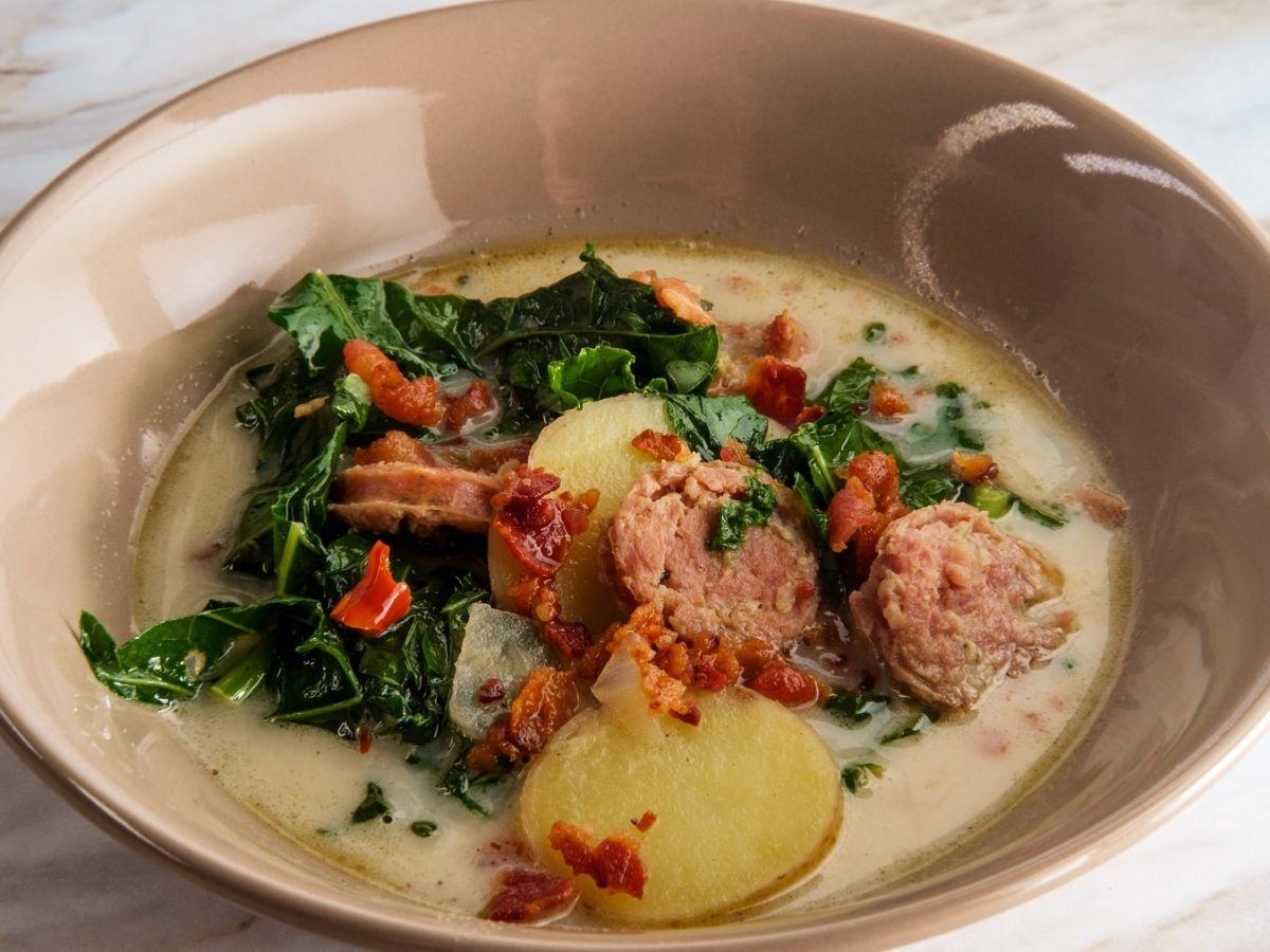 Eine braune Schüssel mit der Grünkohl-Kartoffelsuppe auf einem hellen Marmortisch. Alles von schräg oben fotografiert.