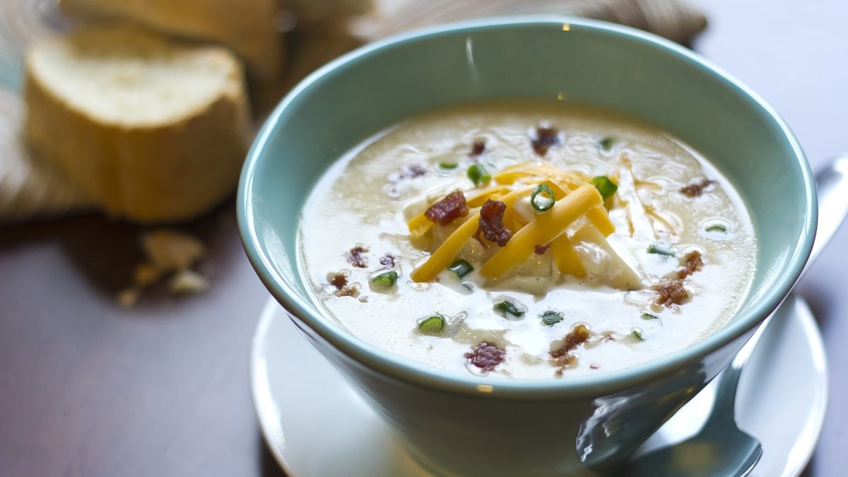 Eine türkise Schüssel mit der Kartoffel-Käse-Suppe auf einem grauen Tisch mit Brot im Hintergrund. Alles von schräg oben fotografiert.