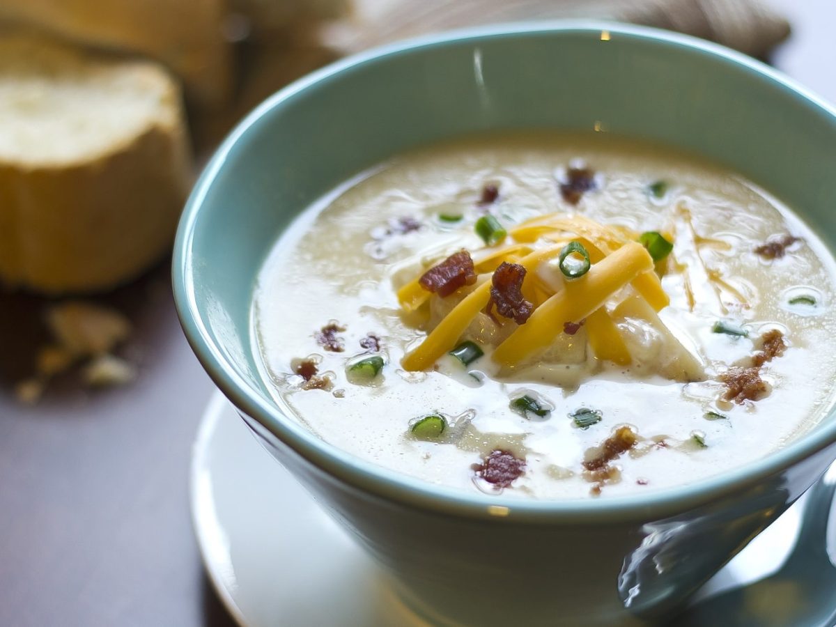 Eine türkise Schüssel mit der Kartoffel-Käse-Suppe auf einem grauen Tisch mit Brot im Hintergrund. Alles von schräg oben fotografiert.