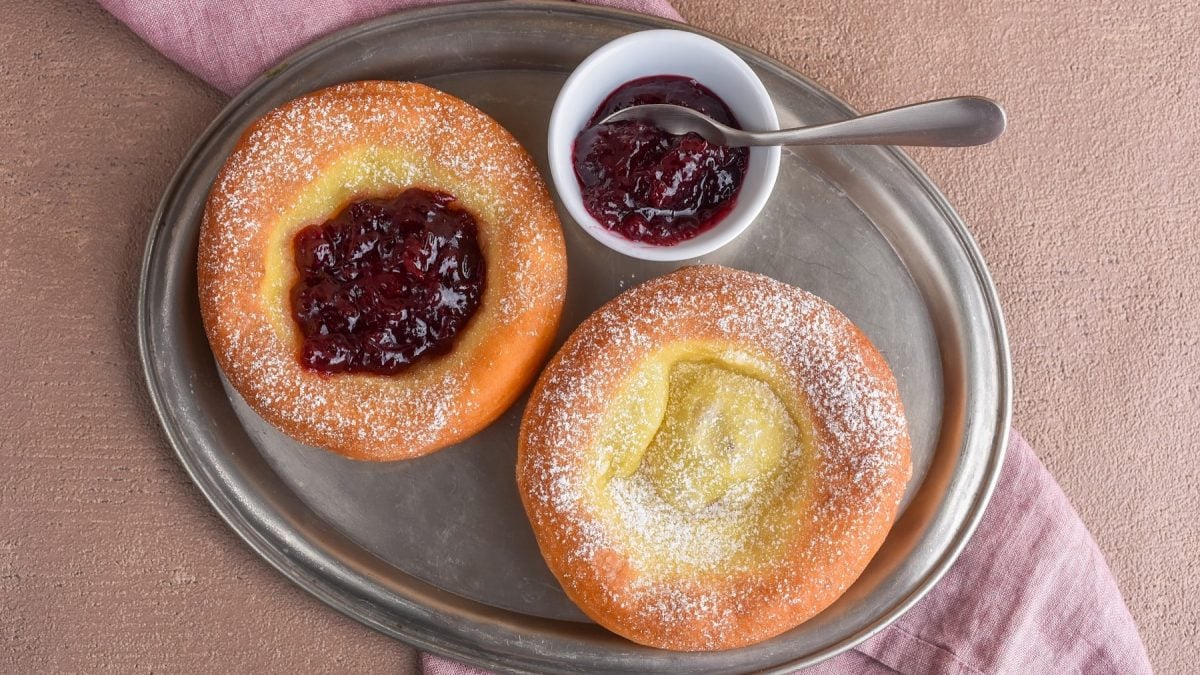 Zwei Stück Tiroler Kiachl auf einem silbernen Servierteller auf einem roten Geschirrtuch. Auf dem Teller ein kleines Schüsselchen mit Preiselbeeren und ein kleiner Löffel. Der Hintergrund ist beige.