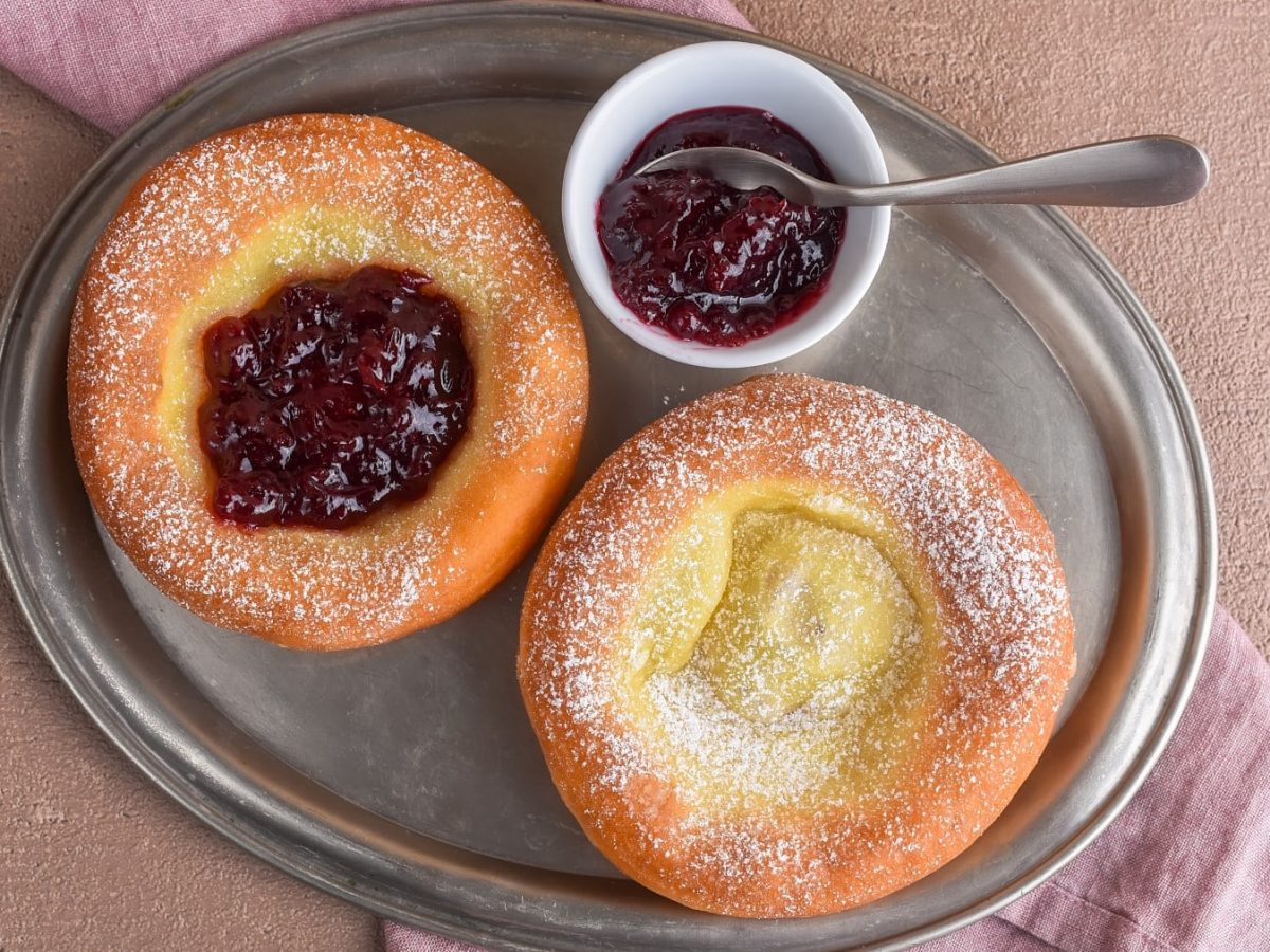 Zwei Stück Tiroler Kiachl auf einem silbernen Servierteller auf einem roten Geschirrtuch. Auf dem Teller ein kleines Schüsselchen mit Preiselbeeren und ein kleiner Löffel. Der Hintergrund ist beige.