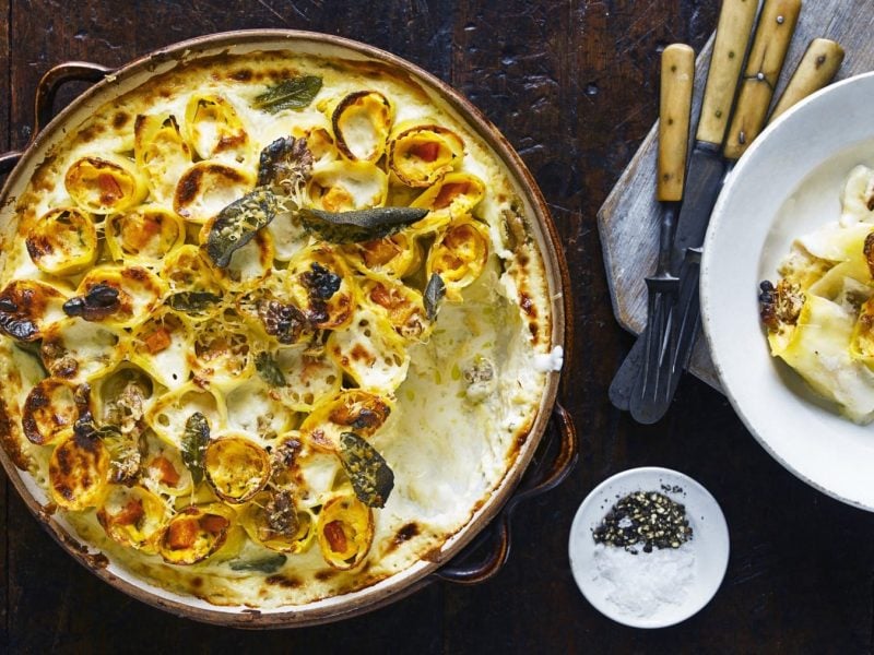 Kürbis-Ricotta-Cannelloni mit Salbei in einer Auflaufform auf einem hölzernen Hintergrund. Rechts davon ein Teller mit einer Portion Cannelloni, Besteck und ein kleines Schüsselchen mit Salz und Pfeffer.