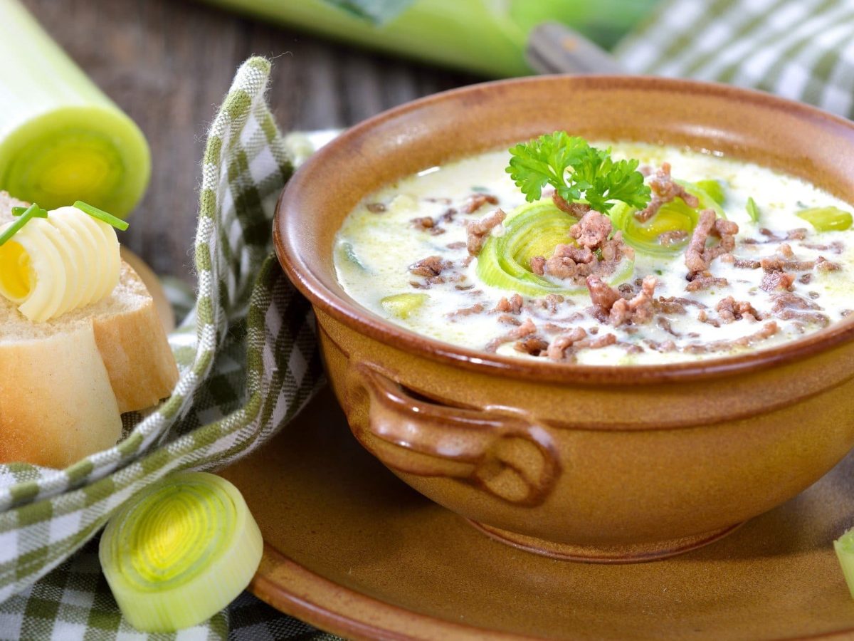 Draufsicht, schräg: Eine Ton-Schüssel mit einer Lauchsuppe mit Hackfleisch. Drum herum liegen Lauchstangen, Lauchringe sowie ein eine Scheibe Weißbrot mit Butter.